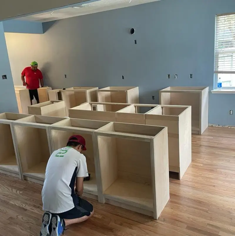 A worker assembling wooden cabinets in a room with unfinished flooring and walls, showcasing the cabinet layout and construction.